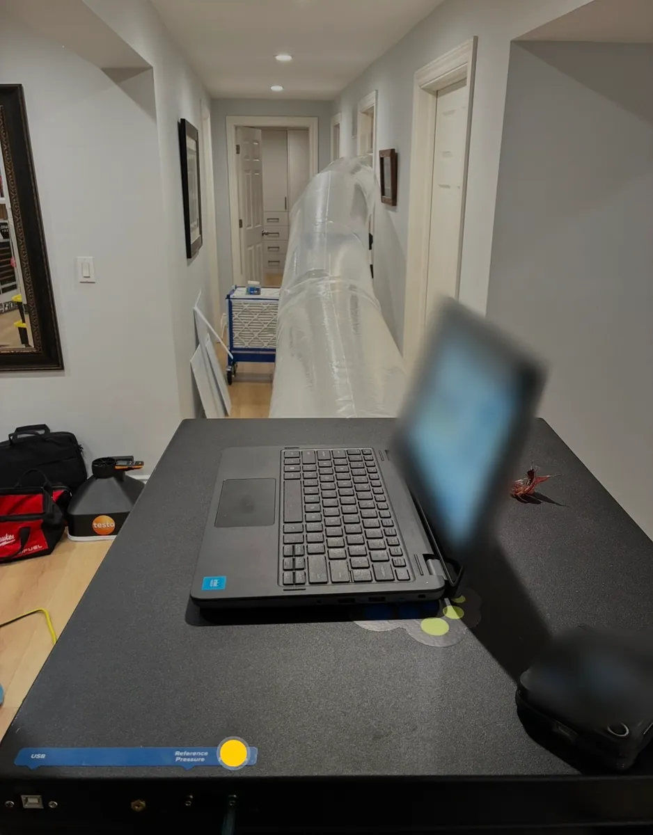 Duct sealing machine with a laptop on top in a hallway, a clear tube running down the hall, a testo instrument and tool bags on the floor