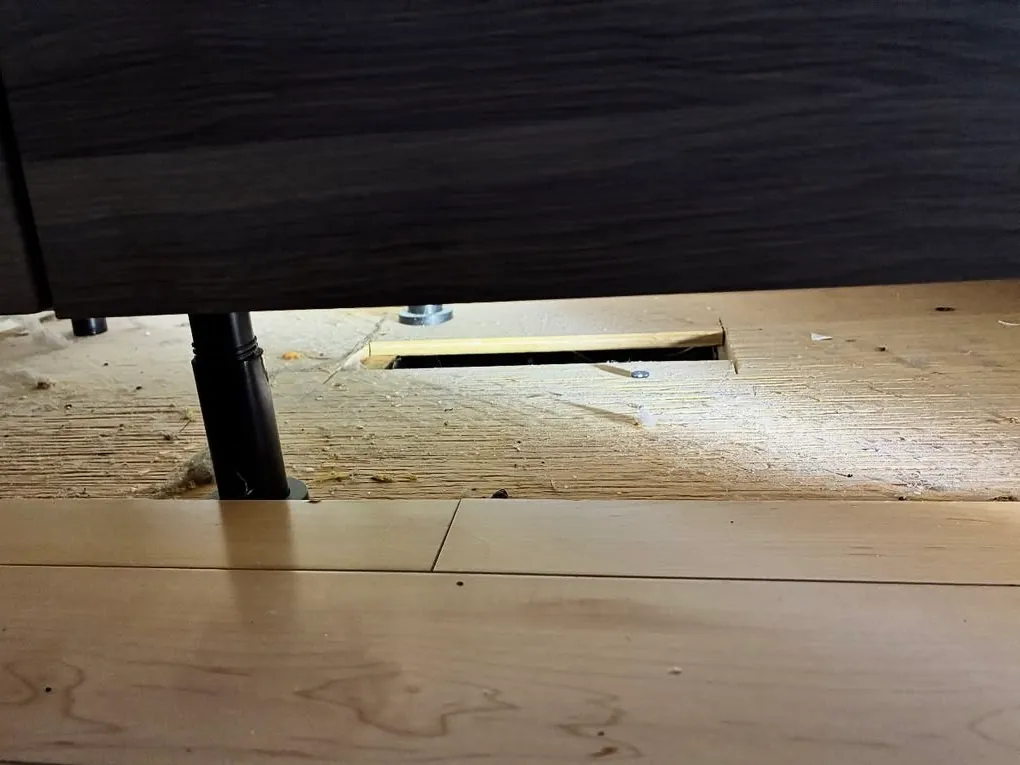 The space under kitchen base cabinets with the kickplate removed, showing a cabinet leg, the subfloor and a register opening in the floor behind it