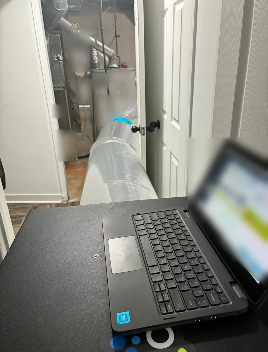 Clear Aeroseal injection tube running from a finished basement into the mechanical room with the furnace and water heater, with a laptop in the foreground