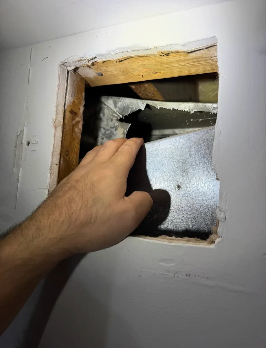 Opening in a drywall wall with a hand reaching into a large torn hole in the metal duct behind it