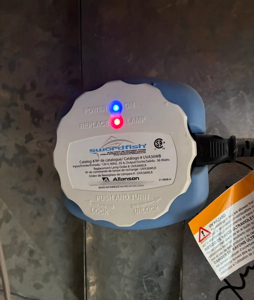 Round white UV air treatment unit with a blue base, mounted on sheet-metal ductwork and plugged in, with an orange UV hazard tag hanging beside it