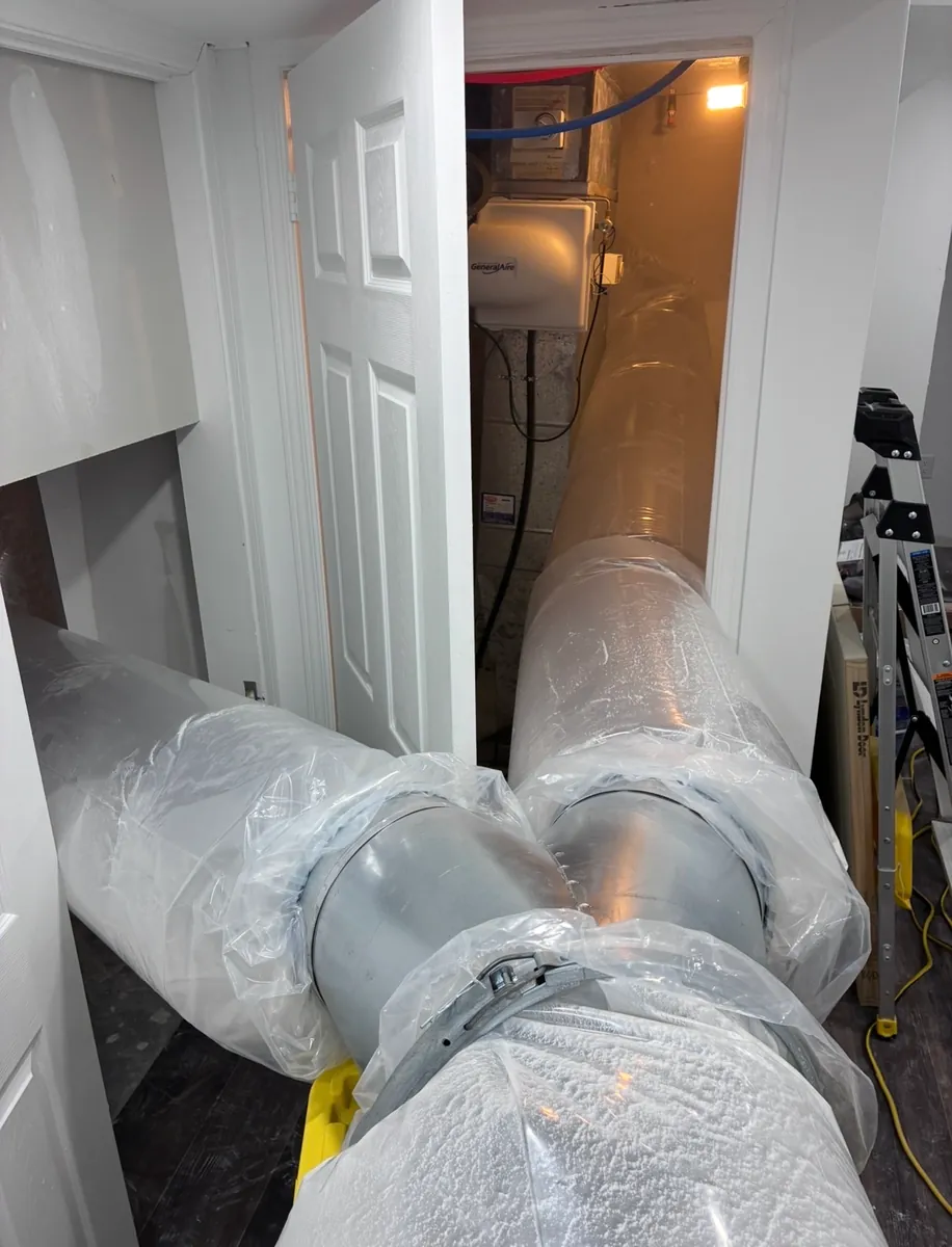Large clear sealing tubes joined together with a metal clamp, running along a hallway floor and through a doorway into a small mechanical room with a humidifier on the wall
