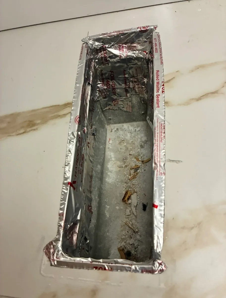 Floor register opening with the register removed, showing the metal boot sealed to the floor opening with foil mastic tape