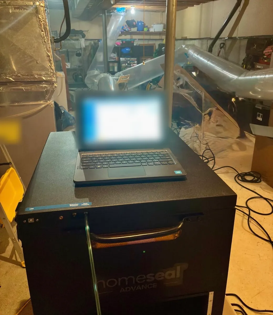 Duct sealing machine with a laptop on top in an unfinished basement, beside the furnace, with large clear tubes running across the room