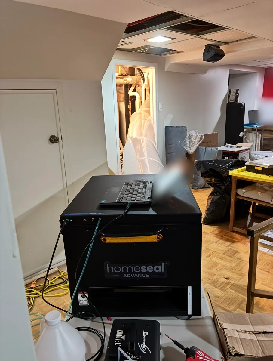 Duct sealing machine with a laptop on top in a finished basement, a clear tube running through a doorway into the mechanical room, and a smoke machine in front
