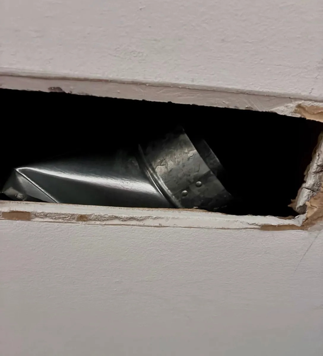 Narrow opening in a basement drywall ceiling showing a metal duct and an elbow fitting that don't line up with each other